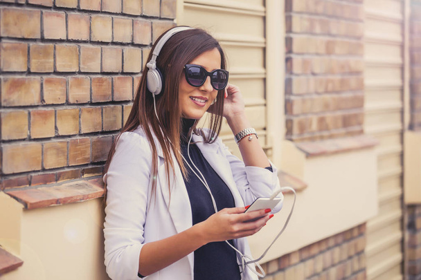 Happy beautiful young woman listening to music via headphones on the street on a sunny day.  Brick Wall Background - Fotoğraf, Görsel