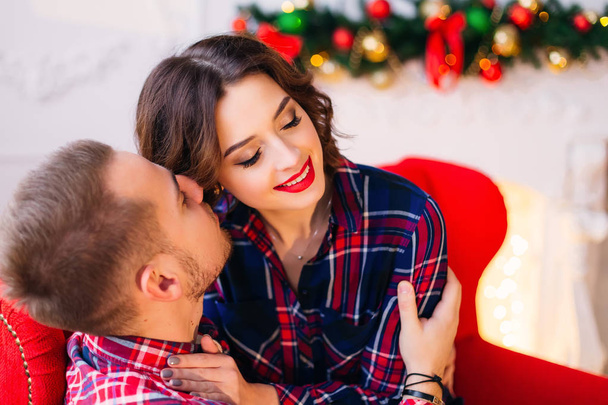 The guy sitting on the couch and hugs his girlfriend and wants to kiss her and she closed her eyes on the background is decorated fireplace - Photo, Image