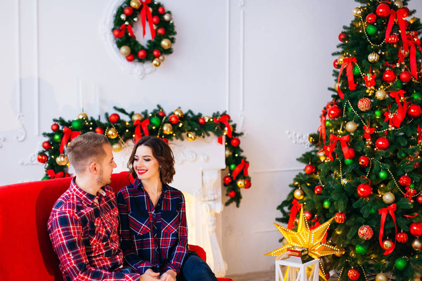 una pareja enamorada sentada en el sofá y mirándose en una habitación con chimenea decorada, árbol de navidad y en la pared una corona de navidad
 - Foto, Imagen