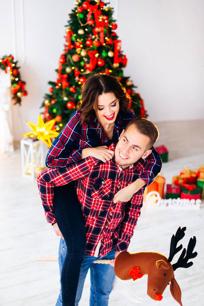 Le gars tient un modèle d'une tête de cerf dans ses mains et la fille a sauté sur le gars et ils ont souri dans le contexte d'un arbre de Noël
 - Photo, image