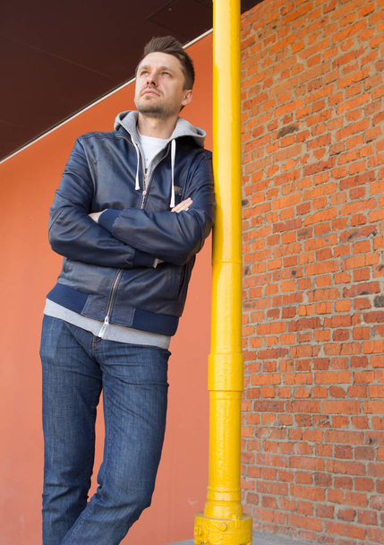 A young attractive man wearing blue jeans and jacket and standing by the gas pipe on red brick wall background. - Foto, immagini