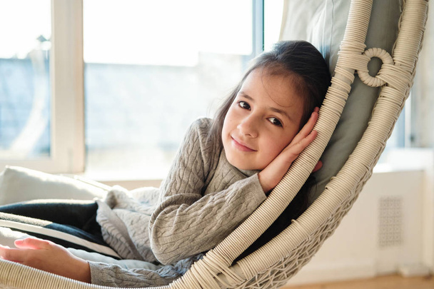 Close up portrait of beautiful young child girl lying at handing chair. Kid relaxing in cozy home, warm soft colors. - Φωτογραφία, εικόνα