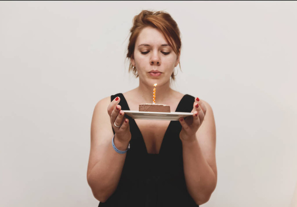A young red-haired woman in a smart black dress holds a cake with candles and blows it out, on a white background. monochrome photo. - Foto, immagini