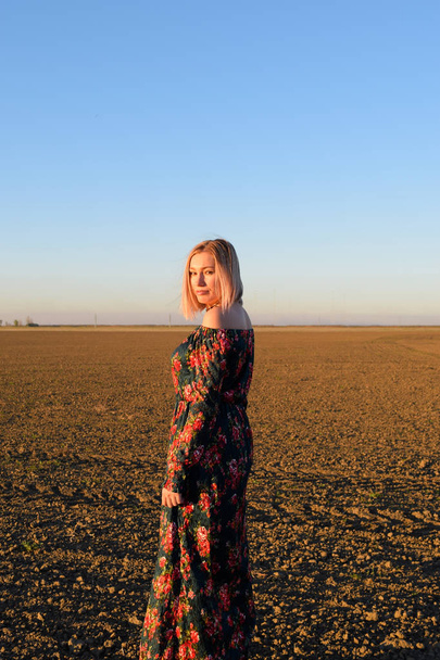 Woman in a plowed field in a red-black dress on a sunset background. - Foto, Imagem