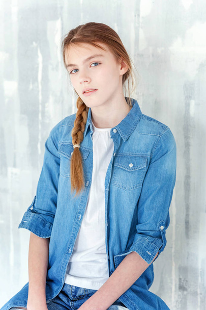 Young happy cute teenage girl in jeans, denim jacket and white T-shirt sitting on chair against grey textured wall background. Casual daily lifestyle. Schoolgirl student education and school concept - Photo, image