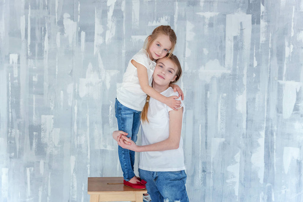 Two happy kids standing against grey textured wall background and embracing. Adorable pretty little girl hugging tight cute teenage girl, showing her love and care. Sisters having fun at home - Φωτογραφία, εικόνα