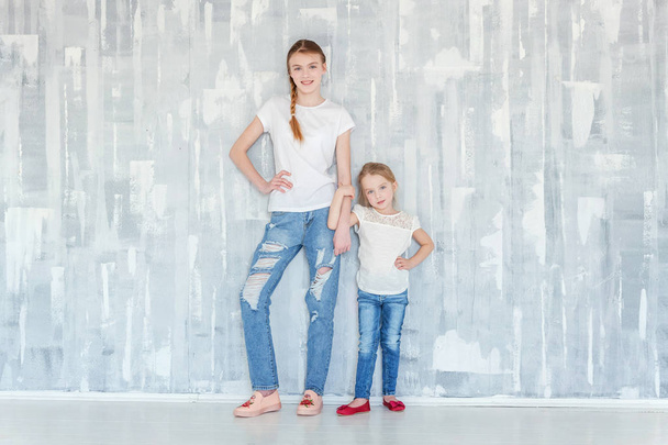 Two happy kids standing against grey textured wall background and embracing. Adorable pretty little girl hugging tight cute teenage girl, showing her love and care. Sisters having fun at home - Zdjęcie, obraz