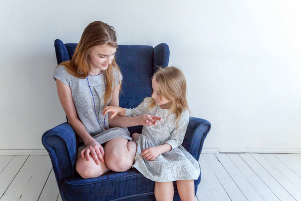 Deux enfants heureux assis sur une chaise bleue confortable moderne relaxant dans le salon blanc à la maison. Adorable jolie petite fille jouant avec une adolescente mignonne, montrant son amour et ses soins. Sœurs s'amuser à la maison
 - Photo, image