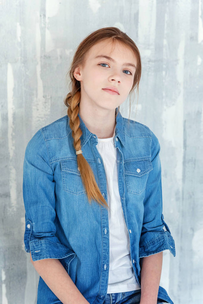 Young happy cute teenage girl in jeans, denim jacket and white T-shirt sitting on chair against grey textured wall background. Casual daily lifestyle. Schoolgirl student education and school concept - Zdjęcie, obraz