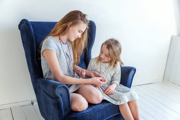 Two happy kids sitting on modern cozy blue chair relaxing in white living room at home. Adorable pretty little girl playing with cute teenage girl, showing her love and care. Sisters having fun at home - Photo, Image