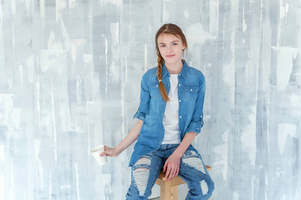 Jeune adolescente mignonne heureuse en jeans, veste en denim et T-shirt blanc assis sur une chaise contre fond de mur texturé gris, tenant une tasse en papier et buvant du café. Style de vie quotidien décontracté
 - Photo, image