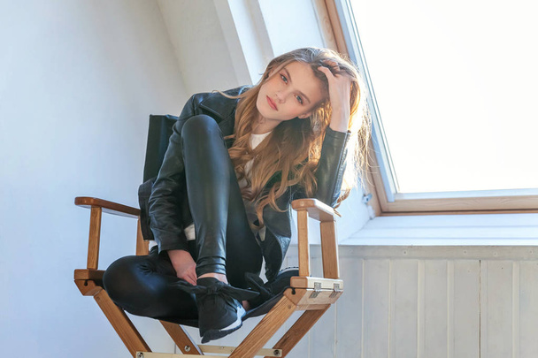 Young teenage girl in leather jacket sitting on modern chair posing in bright light room against white wall background window. Pretty sexy fashion sensual woman dressed in hipster rock style outfit - Foto, imagen