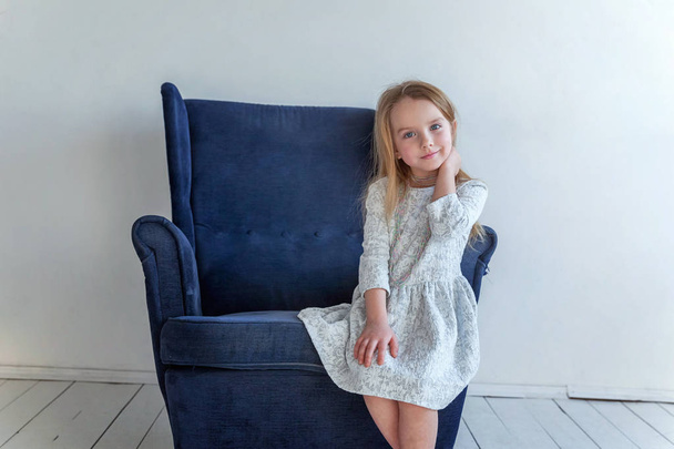 Sweet little girl in grey dress at home sitting on modern cozy blue chair relaxing in white living room - Foto, afbeelding