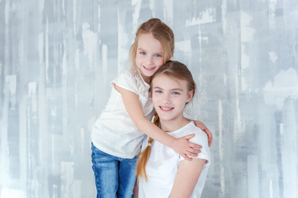 Duas crianças felizes em pé contra fundo de parede texturizado cinza e abraçando. Adorável menina bonita abraçando menina adolescente bonito apertado, mostrando seu amor e cuidado. Irmãs se divertindo em casa
 - Foto, Imagem