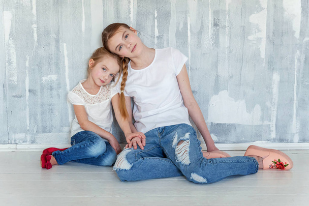 Dwie szczęśliwe dzieci siedząc przed szary teksturowanej tło ściany i ogarnięcie. Urocza dziewczyna dość mało przytulanie napięty cute nastoletnie dziewczyny, pokazując jej miłości i opieki. Siostry, zabawy w domu - Zdjęcie, obraz