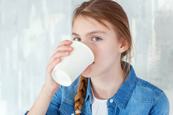 Young happy cute teenage girl in jeans, denim jacket and white T-shirt sitting on chair against grey textured wall background, holding paper cup and drinking coffee. Casual daily lifestyle - Foto, Imagen