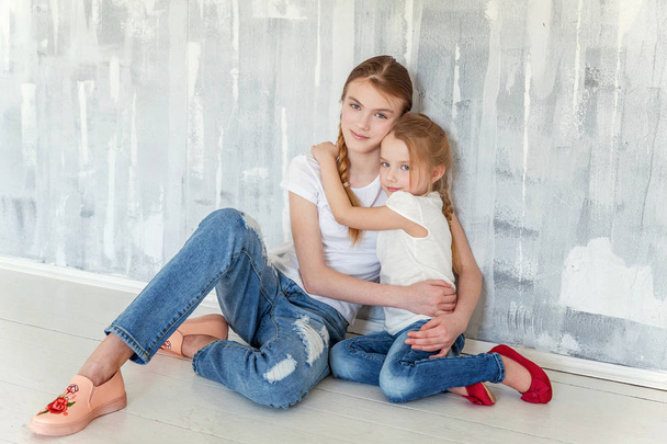Two happy kids sitting against grey textured wall background and embracing. Adorable pretty little girl hugging tight cute teenage girl, showing her love and care. Sisters having fun at home - Photo, Image