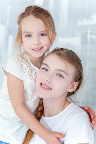 Two happy kids standing against grey textured wall background and embracing. Adorable pretty little girl hugging tight cute teenage girl, showing her love and care. Sisters having fun at home - Fotó, kép