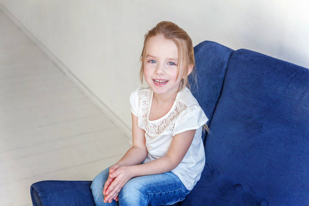 Sweet little girl in jeans and white T-shirt at home sitting on modern cozy blue chair relaxing in white living room - 写真・画像