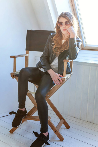 Young teenage girl in leather jacket sitting on modern chair posing in bright light room against white wall background window. Pretty sexy fashion sensual woman dressed in hipster rock style outfit - Photo, Image