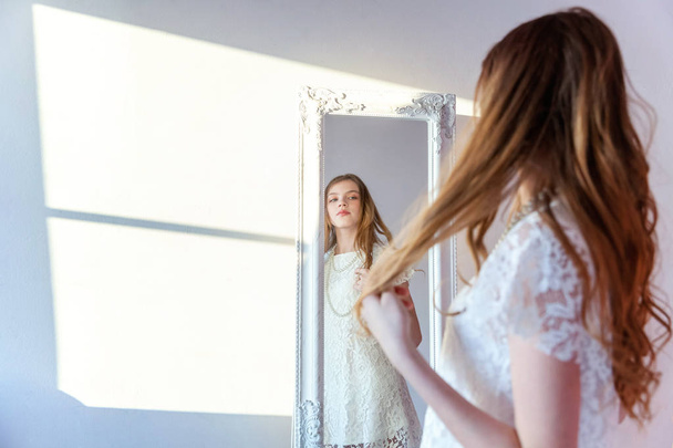 beauty, hygiene, morning and people concept - smiling happy teenage girl looking at reflection in mirror. young positive woman wearing white dress posing in bright light room against white wall - Fotó, kép