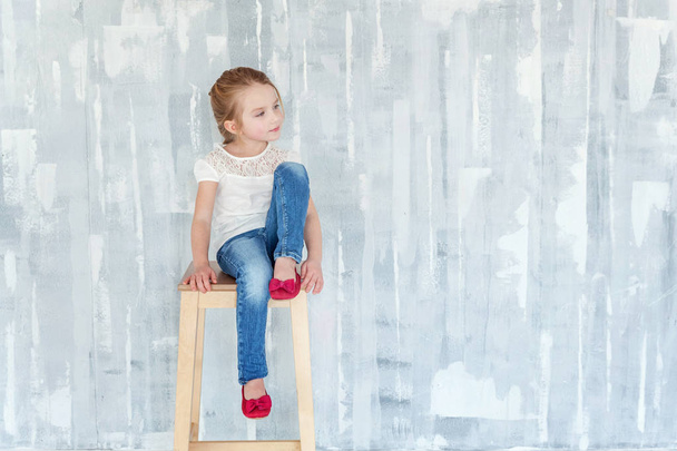 süßes, glückliches kleines Mädchen im weißen T-Shirt sitzt auf einem Stuhl vor grau strukturiertem Wandhintergrund - Foto, Bild