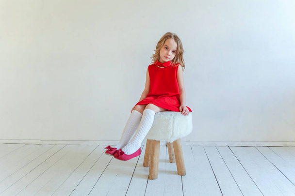 süßes fröhliches kleines Mädchen in rotem Kleid sitzt auf Stuhl vor weißem Wandhintergrund im hellen Raum - Foto, Bild