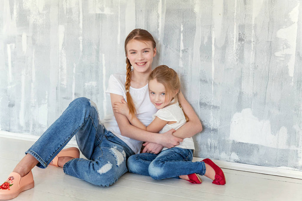 Δύο ευτυχής παιδιά κάθονται σε φόντο γκρι υφή τοίχο και αγκαλιάζει. Αξιολάτρευτο όμορφο κοριτσάκι που αγκαλιάζει σφιχτά χαριτωμένο εφηβικό κορίτσι, δείχνοντας την αγάπη και φροντίδα. Αδελφές έχοντας διασκέδαση στο σπίτι - Φωτογραφία, εικόνα