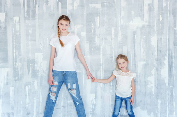 Two happy kids standing against grey textured wall background and embracing. Adorable pretty little girl hugging tight cute teenage girl, showing her love and care. Sisters having fun at home - Photo, Image