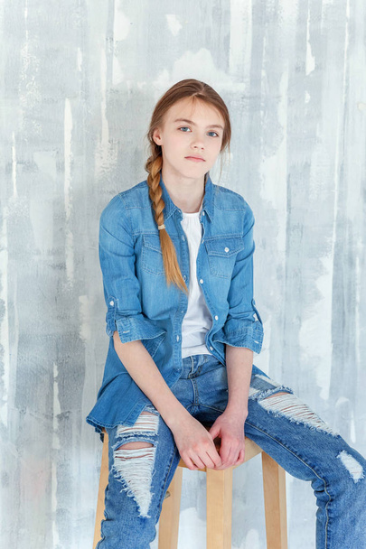 Young happy cute teenage girl in jeans, denim jacket and white T-shirt sitting on chair against grey textured wall background. Casual daily lifestyle. Schoolgirl student education and school concept - Photo, Image