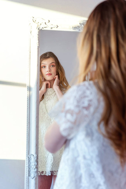 beauty, hygiene, morning and people concept - smiling happy teenage girl looking at reflection in mirror. young positive woman wearing white dress posing in bright light room against white wall - Foto, imagen