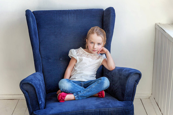 Sweet little girl in jeans and white T-shirt at home sitting on modern cozy blue chair relaxing in white living room - Photo, Image