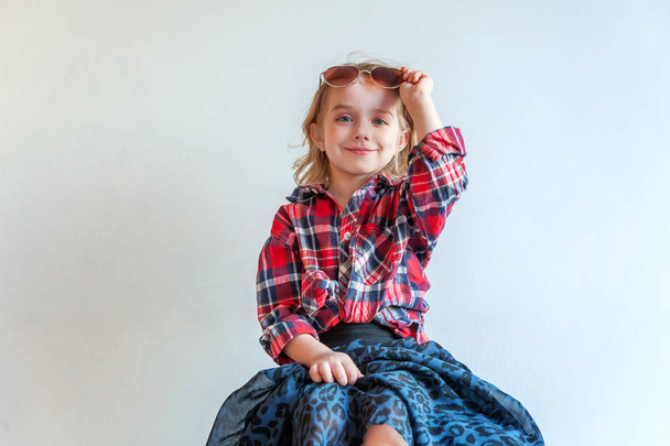 Sweet happy adorable fashion young little girl dressed like hipster hippie gipsy cowboy in sunglasses sitting on modern chair posing in white wall background in light bright room. Lifestyle people trendy concept - Photo, Image
