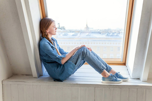 Jovem menina adolescente bonito feliz em jeans, jaqueta jeans e branco T-shirt sentado no peitoril da janela em luz brilhante sala de estar em casa e pensando
 - Foto, Imagem