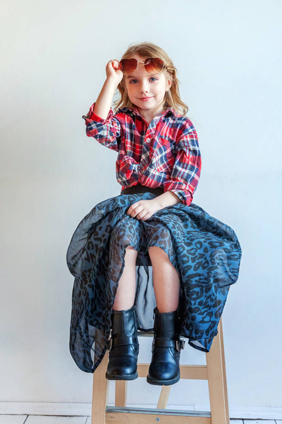 Sweet happy adorable fashion young little girl dressed like hipster hippie gipsy cowboy in sunglasses sitting on modern chair posing in white wall background in light bright room. Lifestyle people trendy concept - Zdjęcie, obraz