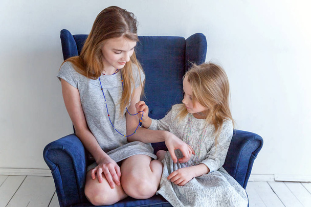 Two happy kids sitting on modern cozy blue chair relaxing in white living room at home. Adorable pretty little girl playing with cute teenage girl, showing her love and care. Sisters having fun at home - Photo, image