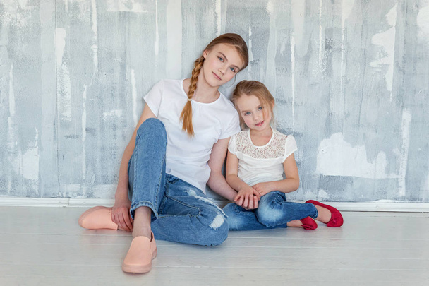 Two happy kids sitting against grey textured wall background and embracing. Adorable pretty little girl hugging tight cute teenage girl, showing her love and care. Sisters having fun at home - Foto, Imagen