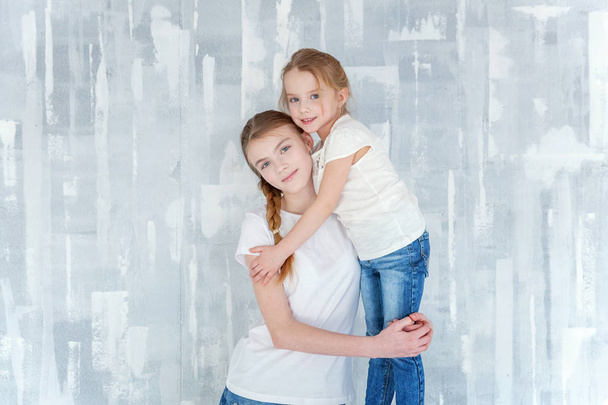 Kaksi onnellista lasta seisoo harmaata kuvioitua seinää vasten ja syleilee. Suloinen kaunis pieni tyttö halaus tiukka söpö teini tyttö, osoittaa hänen rakkautensa ja hoito. Sisarilla on hauskaa kotona
 - Valokuva, kuva