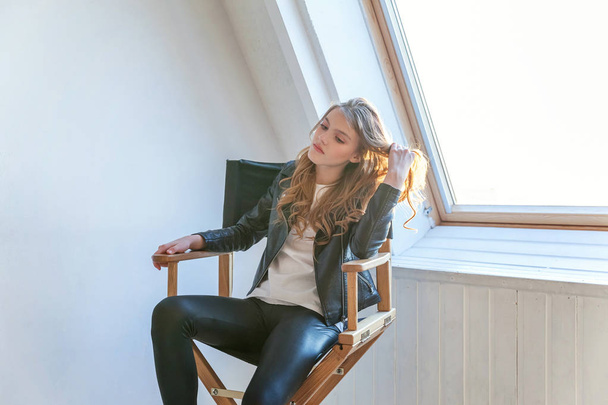 Young teenage girl in leather jacket sitting on modern chair posing in bright light room against white wall background window. Pretty sexy fashion sensual woman dressed in hipster rock style outfit - Zdjęcie, obraz