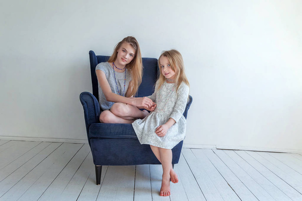 Два счастливых ребенка сидят дома на современном уютном синем стуле, расслабляясь в белой гостиной. Очаровательная маленькая девочка, играющая с симпатичной девочкой-подростком, показывающая свою любовь и заботу. Сестры веселятся дома
 - Фото, изображение