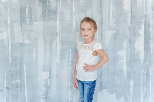 Lief gelukkig klein meisje in lege witte t-shirt staande tegen grijs geweven muur achtergrond - Foto, afbeelding