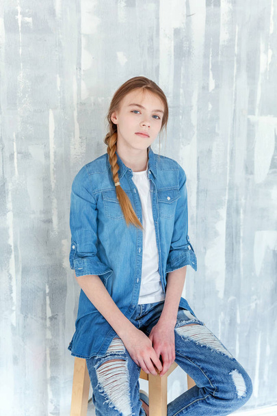 Young happy cute teenage girl in jeans, denim jacket and white T-shirt sitting on chair against grey textured wall background. Casual daily lifestyle. Schoolgirl student education and school concept - 写真・画像