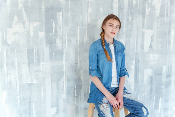 Молодая счастливая девушка-подросток в джинсах, джинсовой куртке и белой футболке, сидящей на стуле на сером текстурированном фоне стен. Обычный повседневный образ жизни. Образование школьниц и школьная концепция
 - Фото, изображение