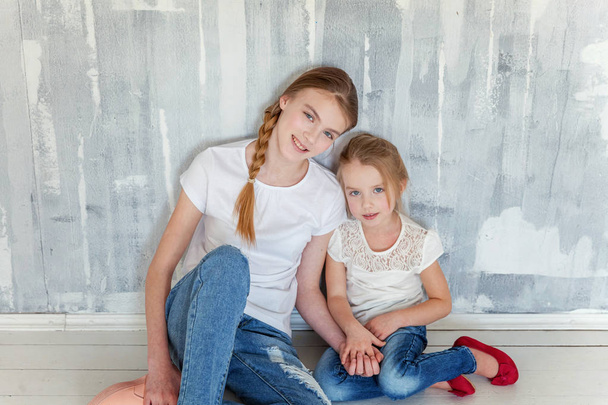 Two happy kids sitting against grey textured wall background and embracing. Adorable pretty little girl hugging tight cute teenage girl, showing her love and care. Sisters having fun at home - Foto, Imagem