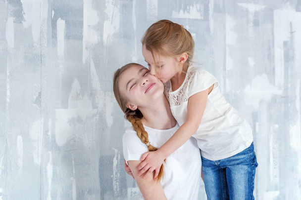 Два счастливых ребенка стоят на сером текстурированном фоне стен и обнимаются. Очаровательная маленькая девочка обнимает обнимающую милую девочку-подростка, проявляет свою любовь и заботу. Сестры веселятся дома
 - Фото, изображение
