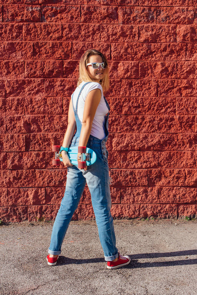 Portrait of a smiling woman dressed in overalls and sunglasses standing with her skateboard next to the red wall. - Φωτογραφία, εικόνα
