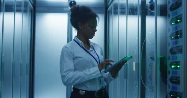 Mulher afro-americana formal usando tablet enquanto trabalhava com rack de servidor no corredor do data center contemporâneo
 - Filmagem, Vídeo