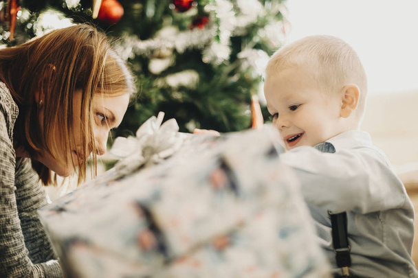 Side näkymä utelias lapsi äiti avaa joululahja laatikko istuu lattialla kotona
 - Valokuva, kuva