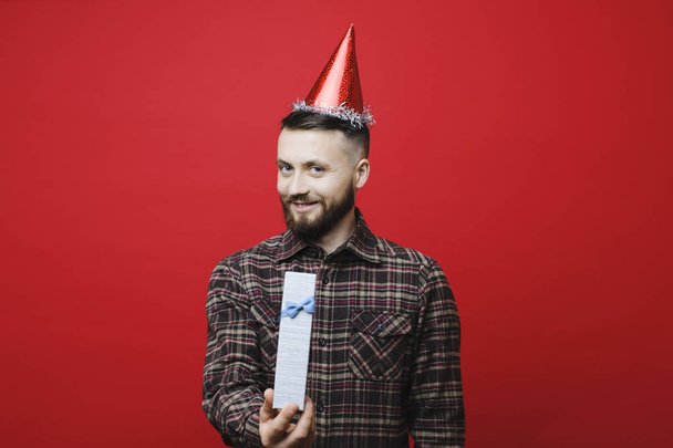 attraktiver bärtiger Typ mit Partyhut, der lächelnd in die Kamera schaut, während er ein schönes Geburtstagsgeschenk macht und auf rotem Hintergrund steht - Foto, Bild