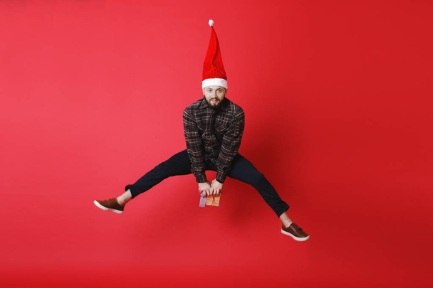 Attractive young man in Christmas hat holding bunch of gifts and looking at camera while jumping on bright red background - Foto, Imagem